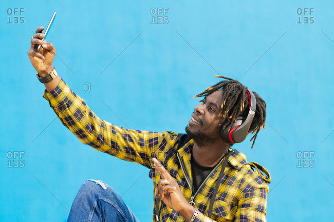 Young black man with headphones taking a selfie with his smart phone with a blue wall in the background, technology and lifestyle concept