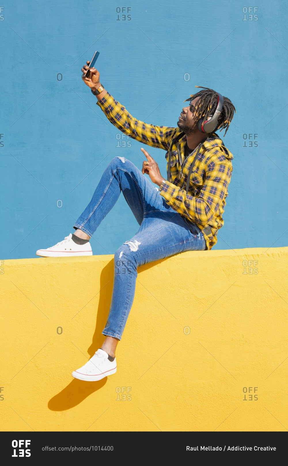 Young black man sitting on a yellow wall with headphones making a video ...