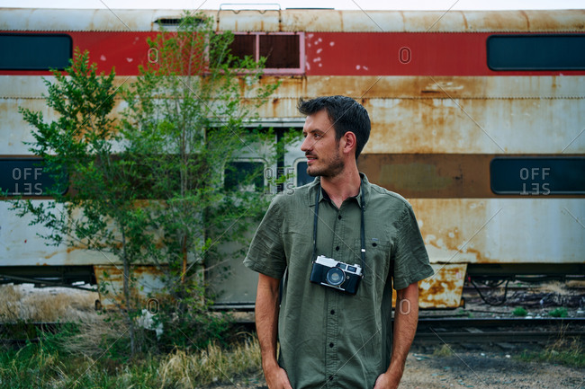 Traveling male photographer with vintage photo camera standing near abandoned rusty train in USA and looking away