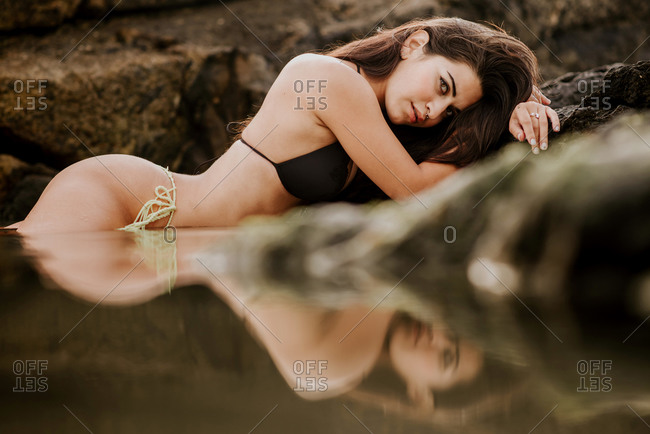 Sensual young female in bikini lying on mud and looking at camera while resting on wet beach