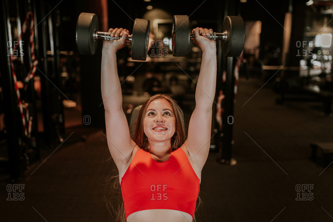 From above happy strong young sportswoman in bright red sportswear doing dumbbell chest press during functional training in modern gym