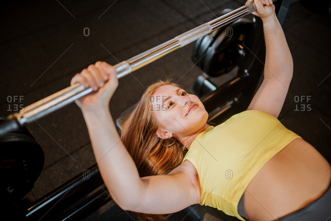 From above of positive young fit female in sportswear performing bench press with barbell during workout in gym