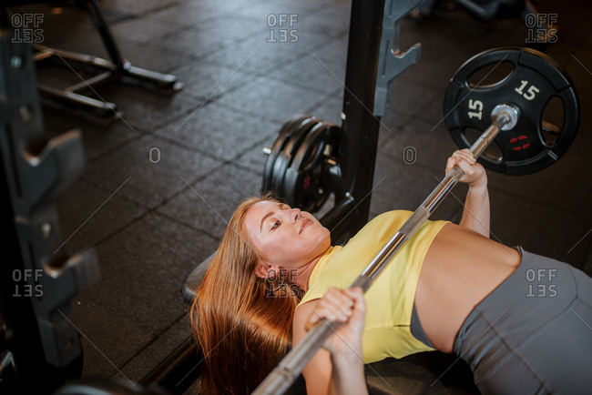From above of positive young fit female in sportswear performing bench press with barbell during workout in gym