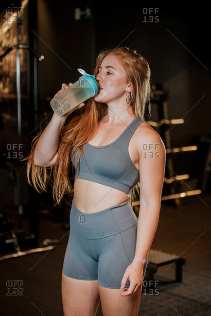 Young fit female in sportswear drinking water from plastic jar while resting after training in gym