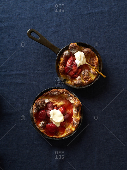 Dutch baby pancakes in various pans with poached strawberries and cream on blue linen background