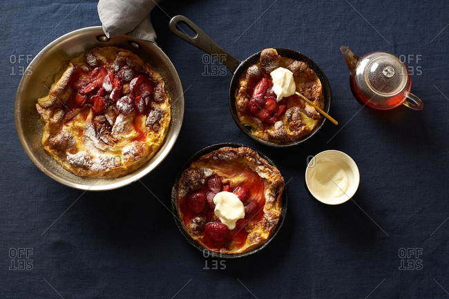 Dutch baby pancakes in various pans with poached strawberries and cream on blue linen background