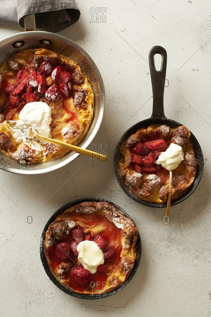 Overhead image of dutch baby pancakes in three different pans with poached strawberries and cream
