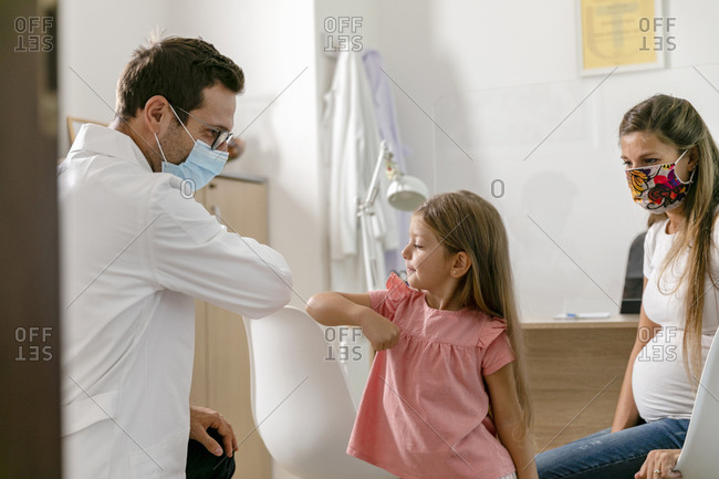 Male medical practitioner in mask bumping elbows with girl near pregnant mother in modern office of hospital during pandemic