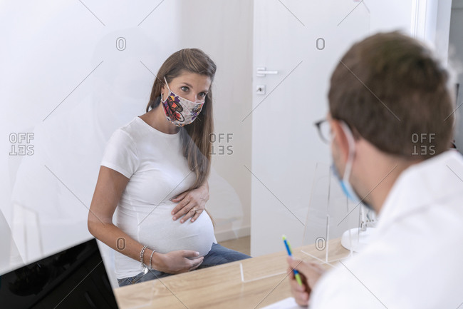 Pregnant female in mask touching belly and speaking with doctor behind glass shield while visiting hospital during epidemic