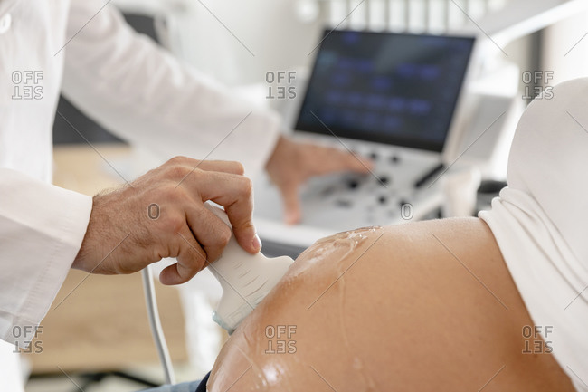 Unrecognizable medical practitioner examining tummy of pregnant woman with ultrasound imaging equipment in clinic