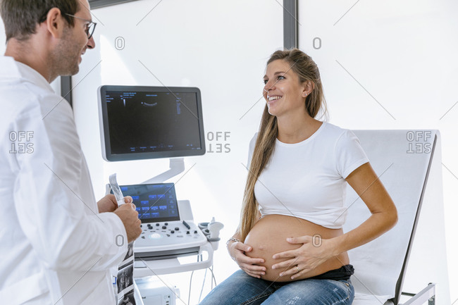 Happy pregnant woman touching tummy and smiling while speaking with medical practitioner after ultrasound imaging procedure