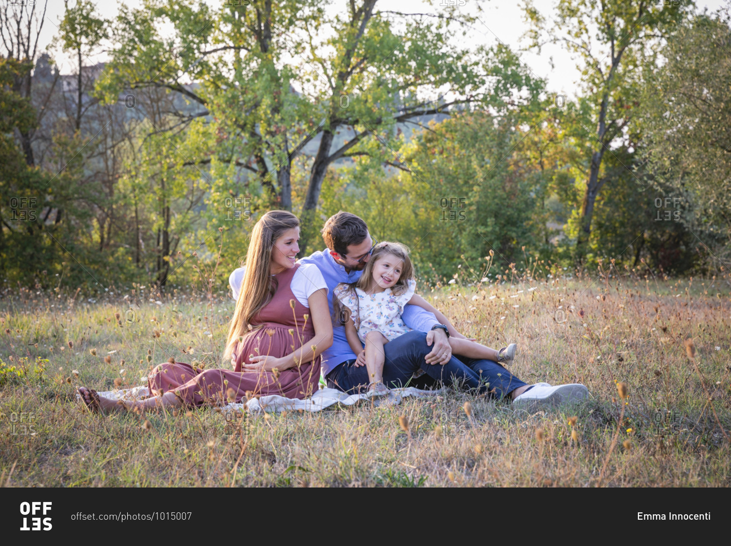 Happy pregnant woman with loving husband and cute little daughter sitting together on grassy meadow during summer weekend in countryside in Italy