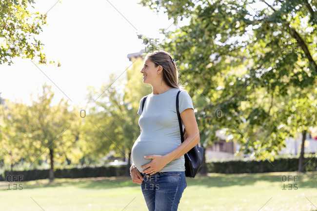 Cheerful young expecting female in casual outfit touching belly and looking away while standing in green park and enjoying sunny summer day