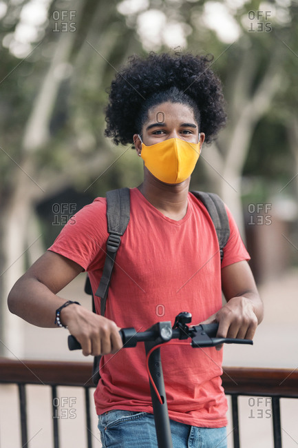 Happy teen black boy wearing face mask due to covid19 and looking at camera while riding an electric scooter