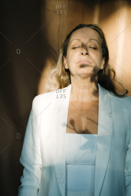 Sunlight on relaxed senior woman standing with eyes closed against curtain