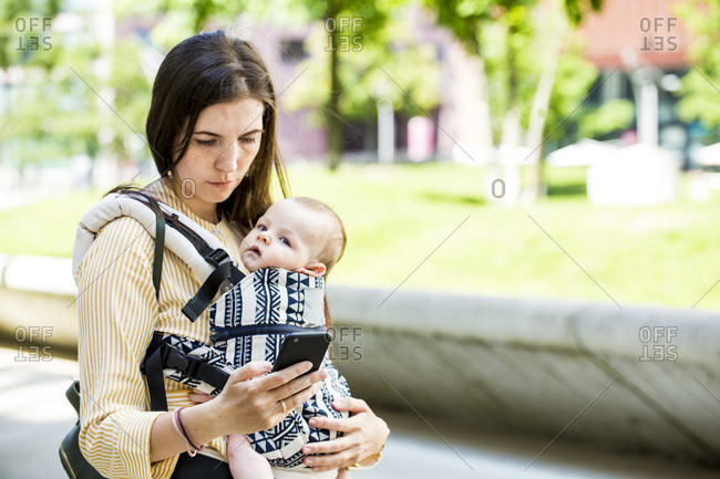 Mother with her baby boy in baby carrier using smartphone in city