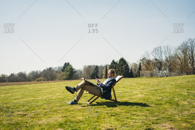 Mature male professional using smart phone while sitting on chair with solar panel at park during sunny day