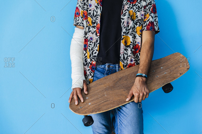 Man with fractured arm holding skateboard while standing against blue background