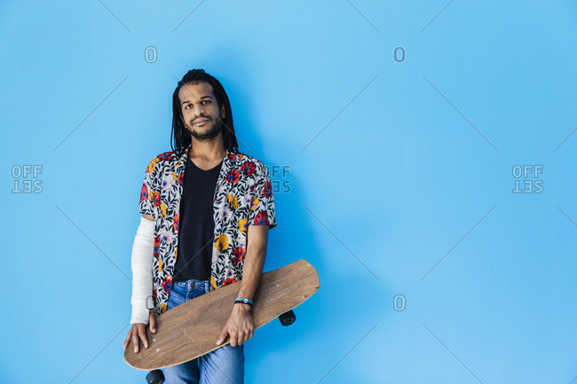Mid adult man with fractured arm holding skateboard against blue background