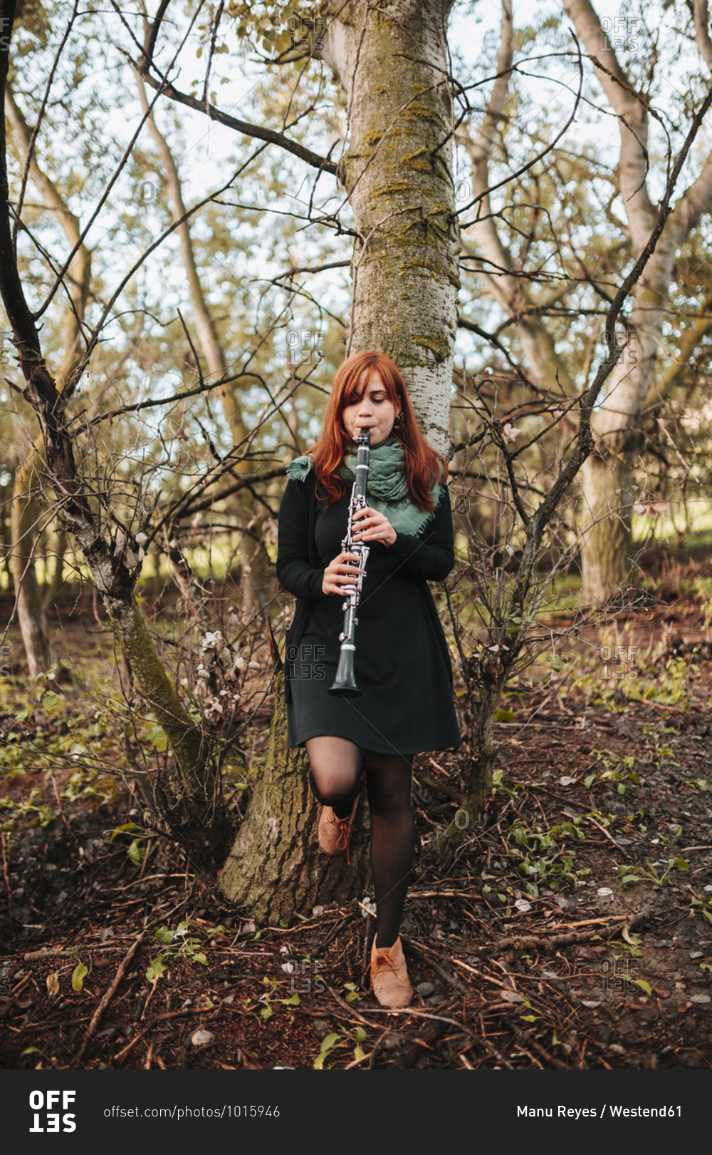 Beautiful young woman practicing clarinet while leaning on bare tree trunk in forest