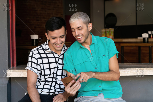 Gay man pointing while sharing mobile phone with friend while sitting restaurant counter