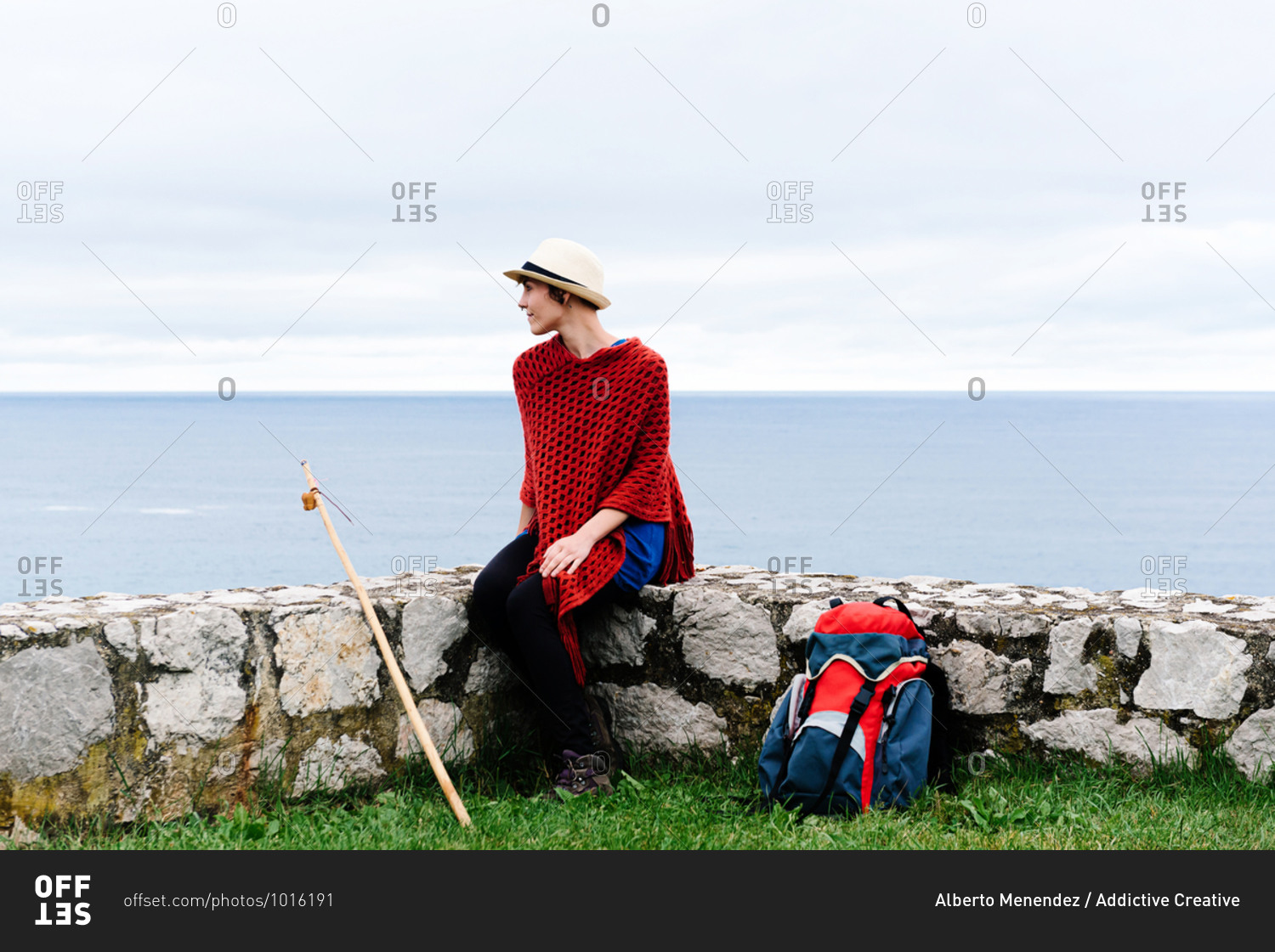 Happy young female backpacker in stylish clothes sitting on stone border against sea bay and looking away while resting during Camino de Santiago route in Spain