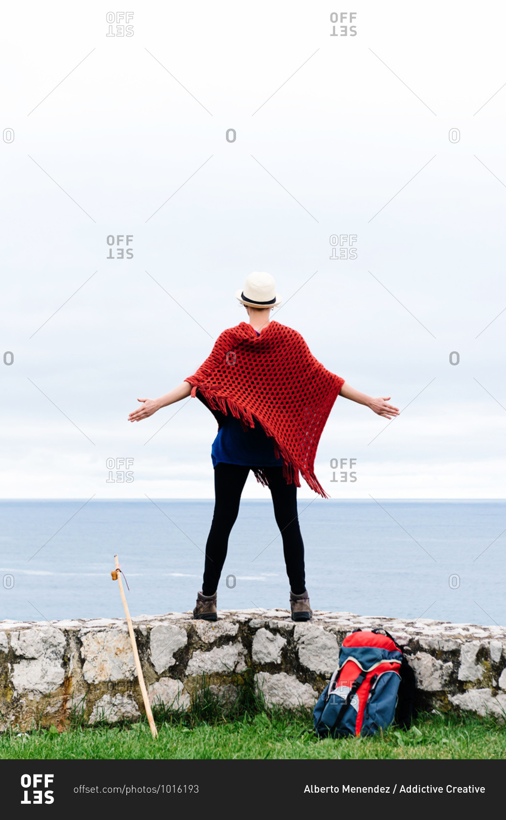 Full body back view of unrecognizable female hiker with backpack and trekking stick standing with arms raised on stone border against gray cloudy sky and enjoying freedom during pilgrimage through Spain