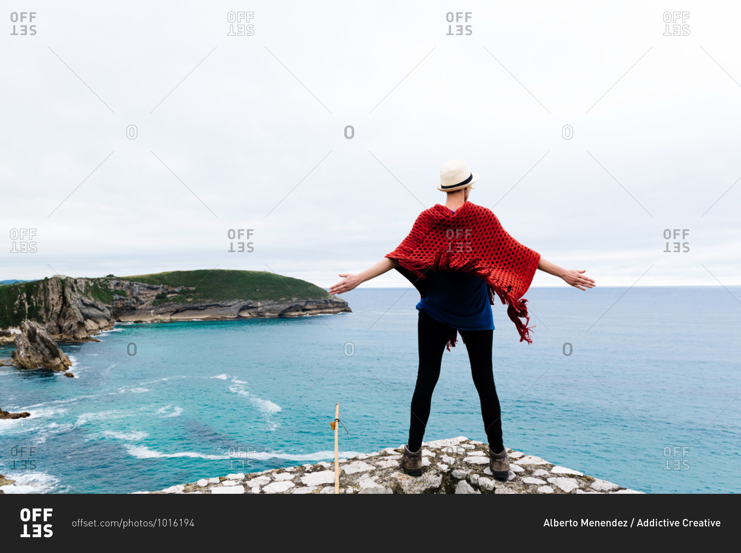 Full body back view of unrecognizable female hiker with backpack and trekking stick standing with arms raised on stone border against gray cloudy sky and enjoying freedom during pilgrimage through Spain