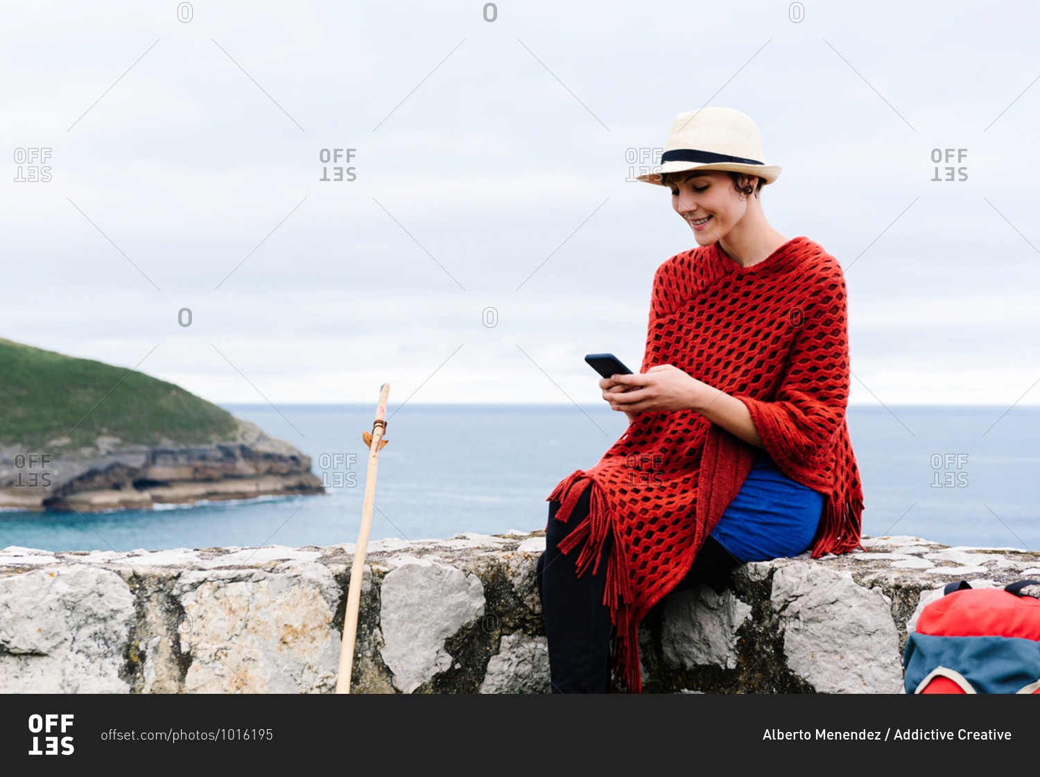Young female backpacker with trekking stick sitting on stone border near sea and browsing smartphone while resting and checking route during pilgrimage through Spain
