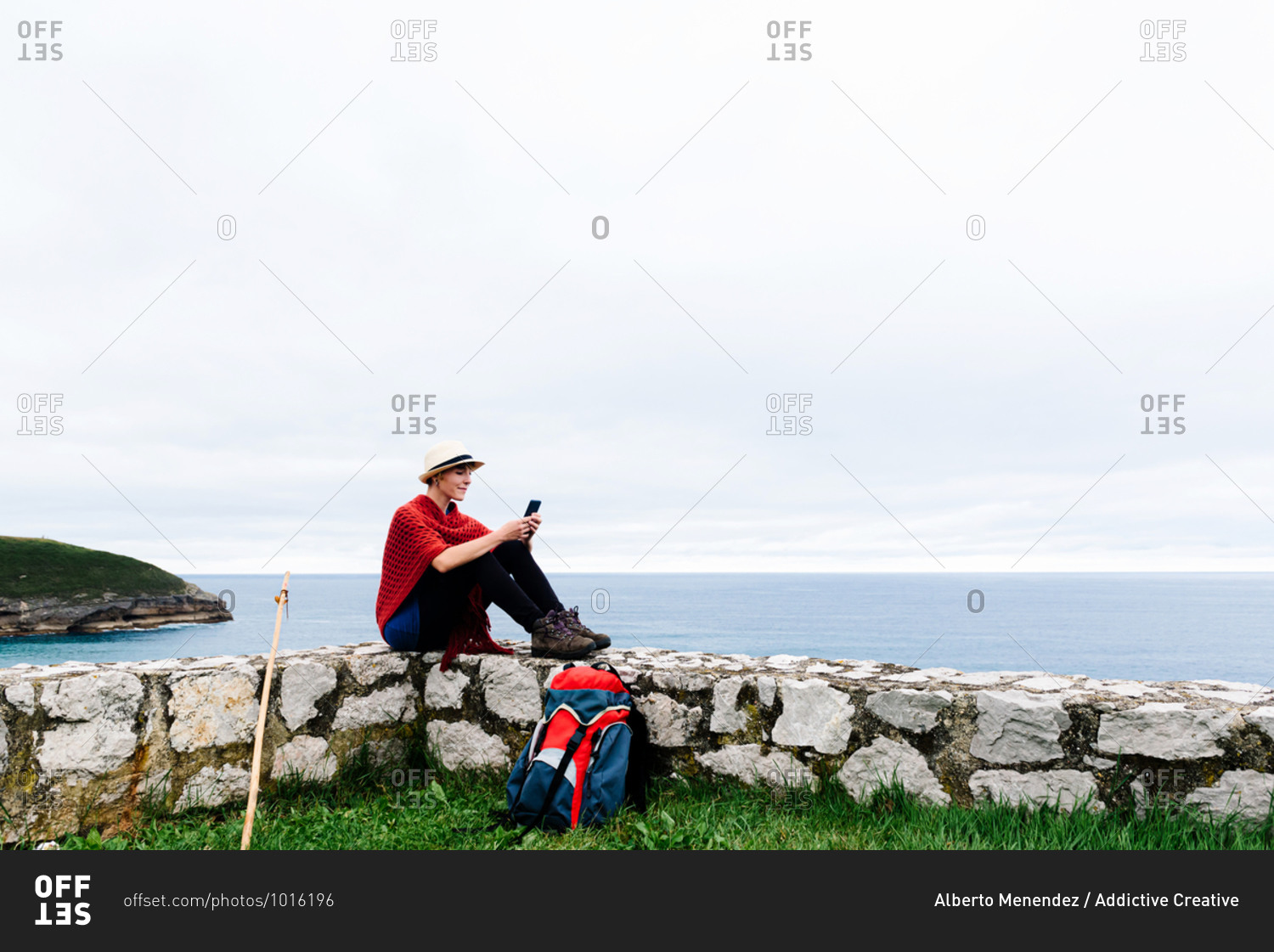 Full length side view of young female backpacker with trekking stick sitting on stone border near sea and browsing smartphone while resting and checking route during pilgrimage through Spain