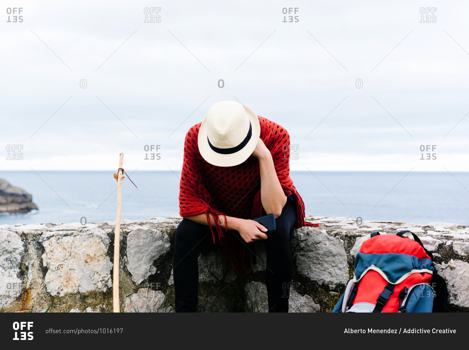 Unrecognizable sad lost female backpacker with trekking stick sitting on stone border near sea and browsing smartphone while resting and checking route during pilgrimage through Spain