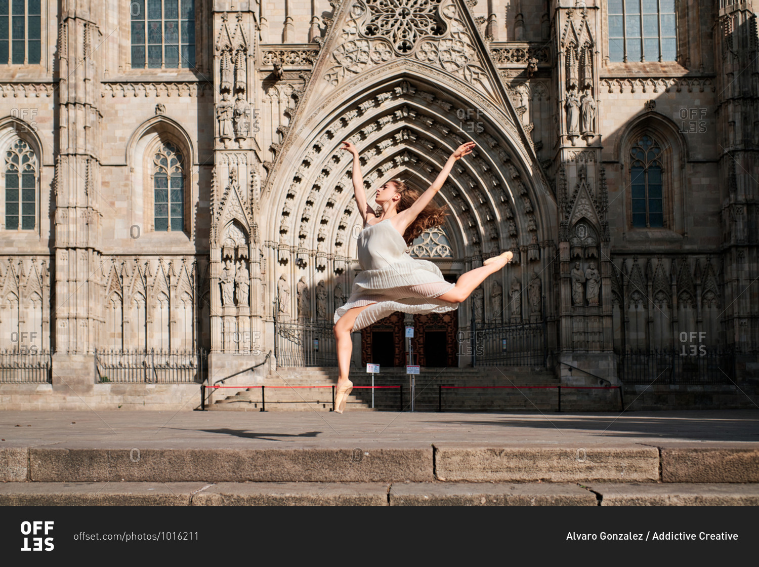 Full body of young female ballet dancer performing sensual dance moves and jumping with arms raised against ornamental stone building with Gothic architecture
