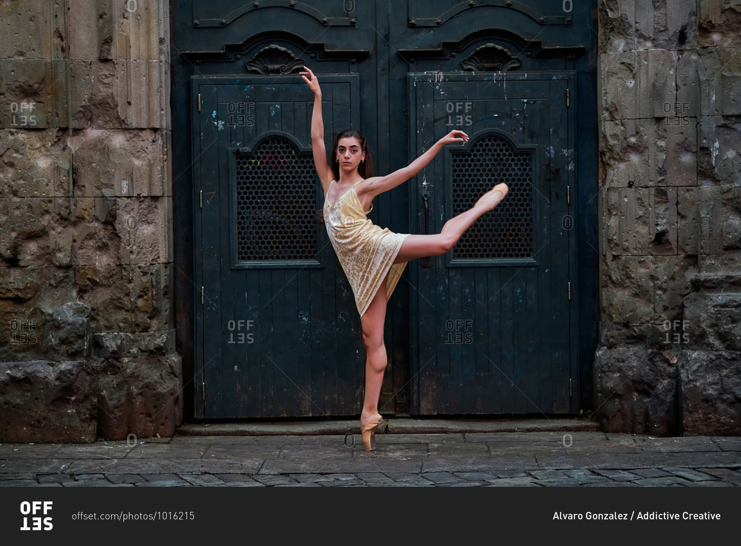 Full body side view of slim young ballerina in dress and pointe shoes performing graceful ballet pose against weathered door of aged stone building on street of old city