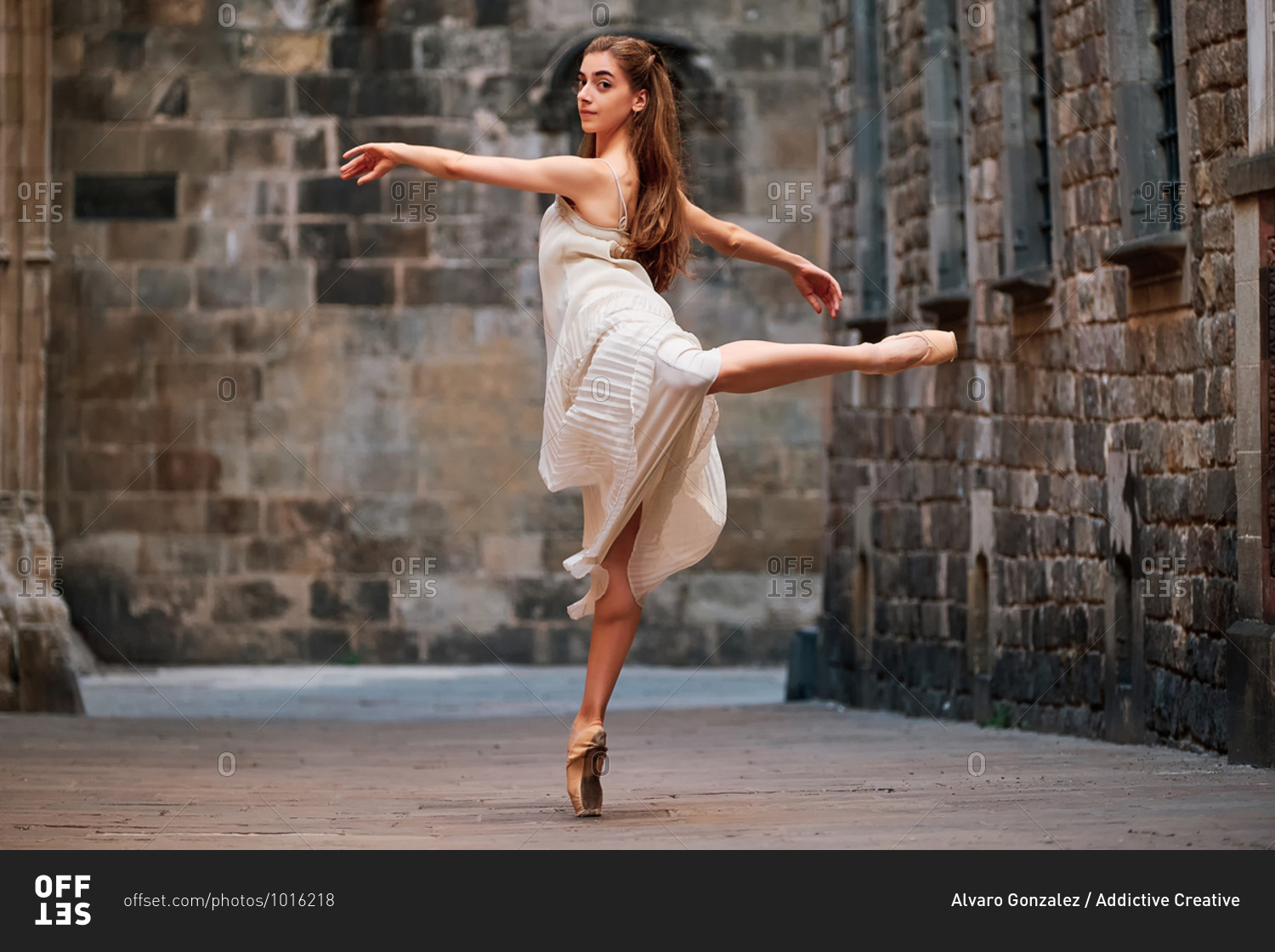 Full body side view of slim young ballerina in dress and pointe shoes performing graceful ballet pose against weathered wall of aged stone building on street of old city