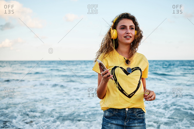 Low angle of optimistic adolescent female in yellow shirt listening to music with smartphone and headphones while resting near waving sea during summer holidays