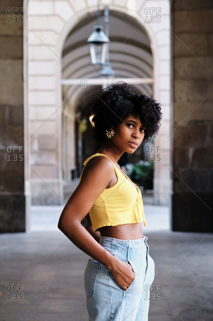 Side view of serious millennial African American female with curly hair and earrings wearing yellow crop top and blue jeans looking at camera while standing against old stone building on city street