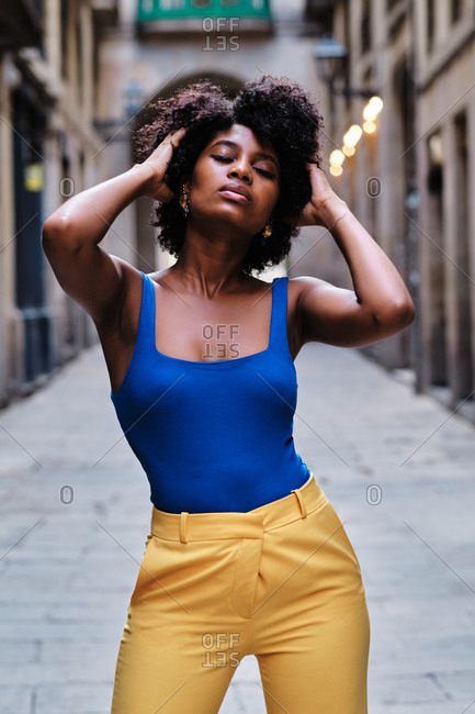 Young slim African American female with curly hair dressed in stylish yellow pants and blue top standing on narrow paved street in old city district touching hair with eyes closed