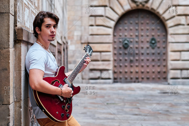 Side view of young male guitarist playing musical instrument on old city street and looking at camera