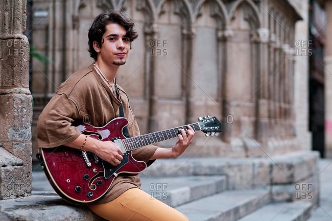 Side view of young male guitarist playing musical instrument on old city street and looking at camera