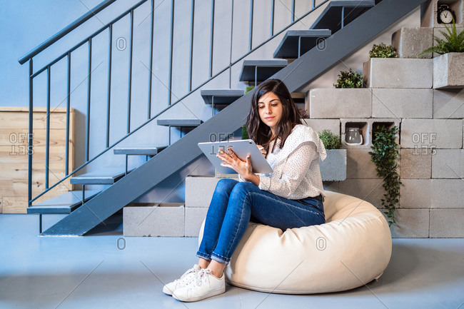 Full length of focused young female designer in casual clothes sitting on comfortable pouf and using graphic tablet while working in contemporary creative workspace