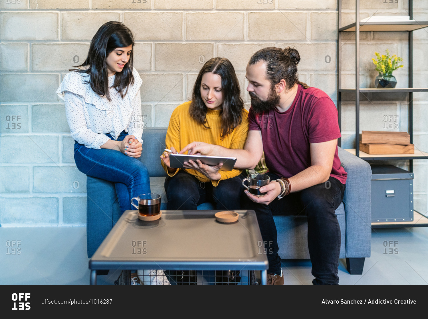 Group of creative millennial designers drinking coffee and discussing design for new project on graphic tablet while gathering in modern loft coworking space