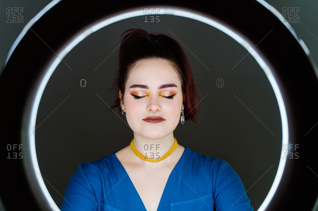 Female with professional makeup and in stylish wear standing in studio behind ring circle lamp on dark background with eyes closed
