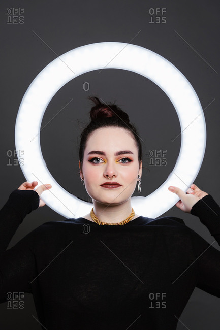 Delighted female with professional makeup and in stylish wear standing in studio holding circle lamp on grey background and looking at camera