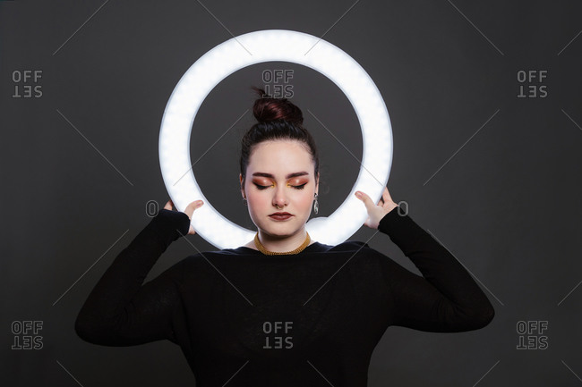 Female with professional makeup and in stylish wear standing in studio holding circle lamp on grey background with eyes closed