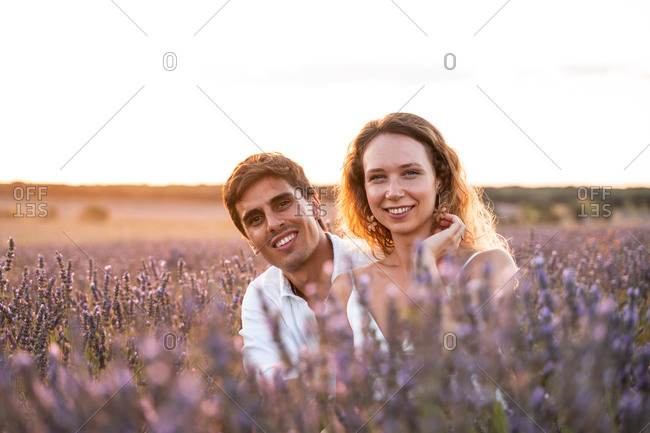 Happy young man and woman sitting together among lavender flowers and enjoying summer evening together during romantic holidays in countryside