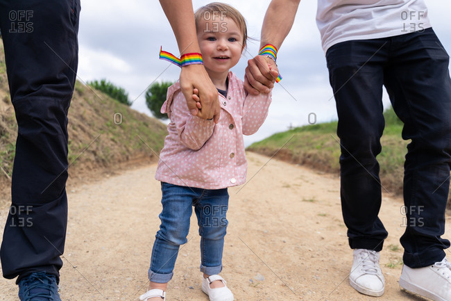 Cute little girl in casual wear holding hands of crop unrecognizable parents with rainbow bracelets while walking together on narrow path in summer countryside