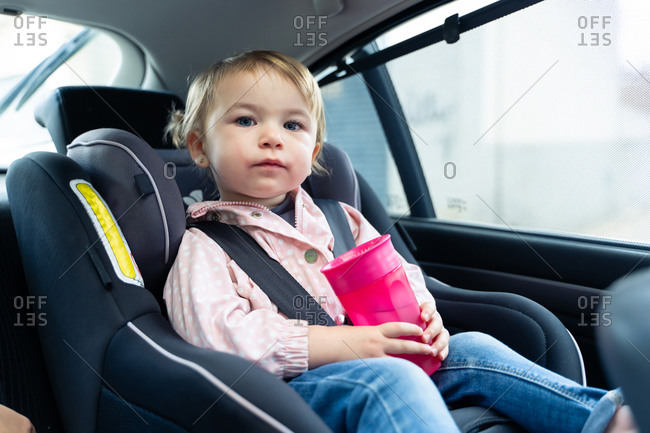 Girl sitting in safety seat with belt fastened and drinking water from plastic cup during trip