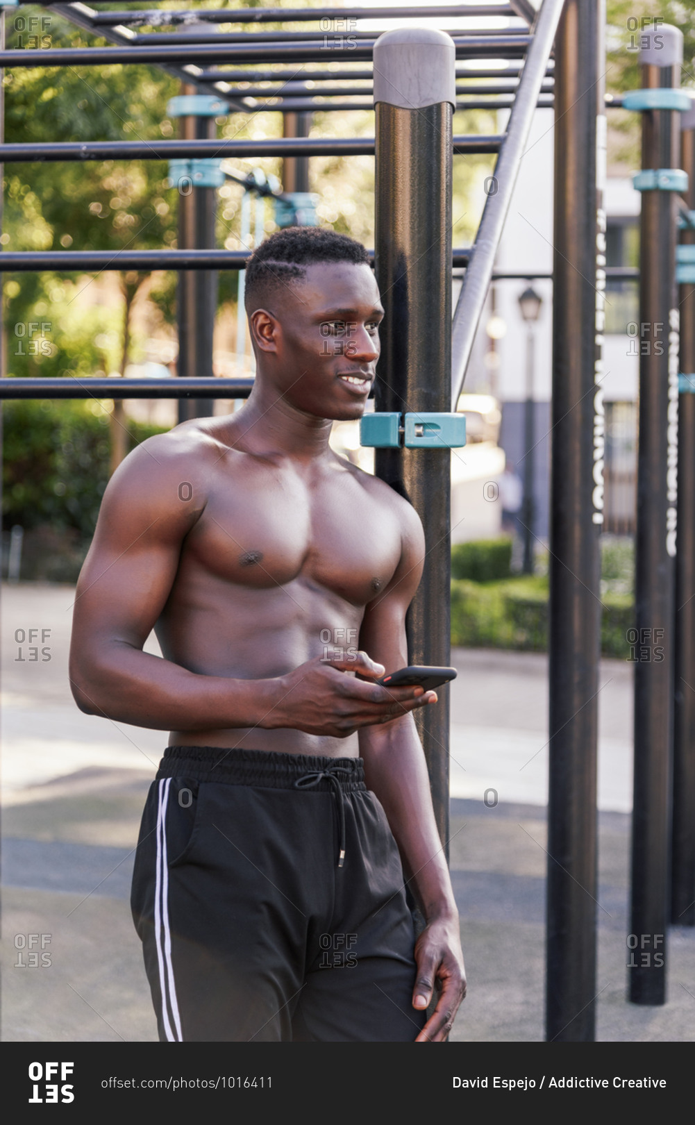 Shirtless African American sportsman leaning on metal bars on sports ground and chatting on social media via cellphone after workout while looking down