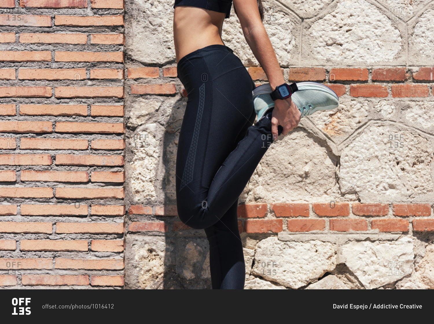 Crop female athlete in leggings standing near brick wall on street and warming up legs during workout