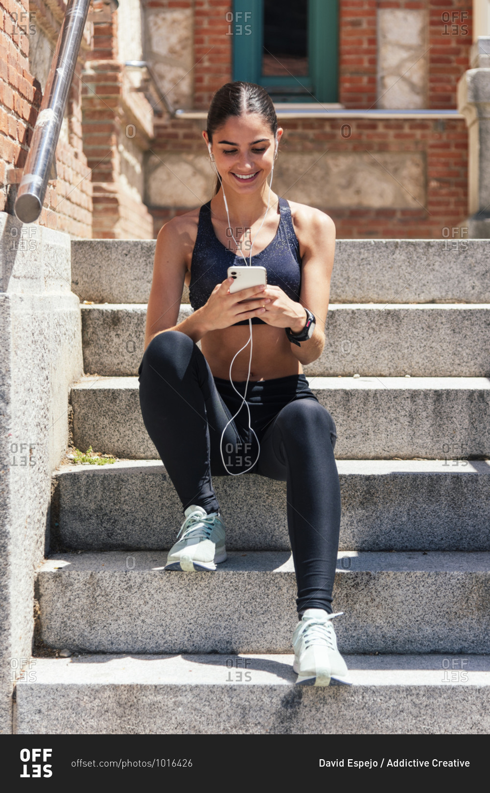 Cheerful female athlete in earphones sitting on steps in street and choosing songs while using smartphone before training